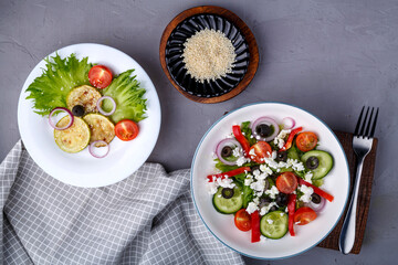 White plate with grilled zucchini with cherry tomatoes on lettuce leaves and greek salad on a concrete background near sesame seeds.