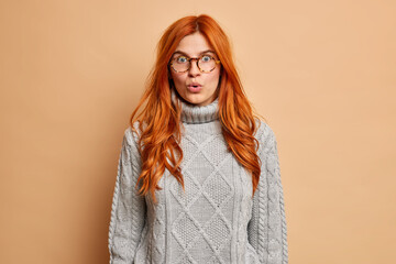 Wall Mural - Photo of wondered ginger woman stares bugged eyes holds breath stands speechless anxious to hear unbelievable news wears transparent glasses and grey knitted sweater poses over beige studio wall