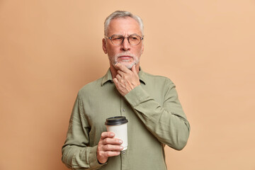 Wall Mural - Pensive wrinkled man stands in thoughtful pose rubs chin and tries to make his mind contemplates about something while has coffee break wears optical glasses and formal shirt. Contemplative pensioner