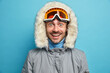 Cheerful unshaven man with overjoyed expression smiles broadly wears ski goggles winter jacket with hood enjoys extreme winter sport poses against blue background. Snowboarding hobby concept