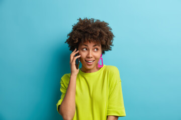 Wall Mural - Pretty cheerful woman with curly hair has telephone conversation talks via mobile phone has glad expression dressed in casual t shirt isolated over blue background. Afro American girl calls friend