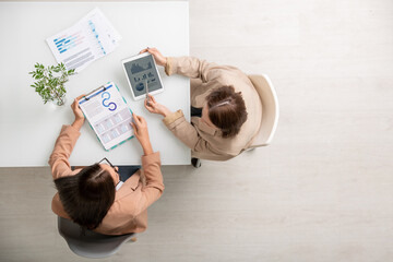 Poster - Overview of two young elegant businesswomen sitting by desk, analyzing data