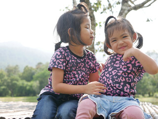 Two little girls, sisters, 3 and 2 years old, kissing on a bamboo bench in the evening sunlight - sisters bond, love, and friendship