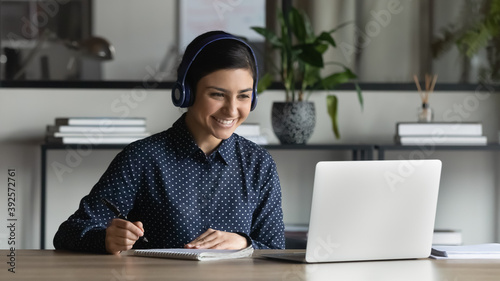 Obraz na płótnie Smiling Indian businesswoman intern wearing headphones watching webinar, writing notes, listening to lecture, happy young female employee student looking at computer screen, studying online