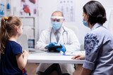 Fototapeta Na drzwi - Pediatrician wearing face mask holding tablet pc while consulting child during clobal pandemic with coronavirus. Pediatrician specialist with protection mask providing health care service.