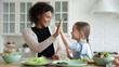 Smiling African American mother and adopted Caucasian daughter giving high five close up, sitting at table in kitchen, celebrating success, multiracial parent and child cooking salad together