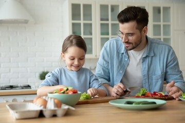 Wall Mural - Caring father and little daughter chopping fresh vegetables on board, cooking salad together, sitting at wooden table in kitchen at home, preparing meal, family enjoying leisure time