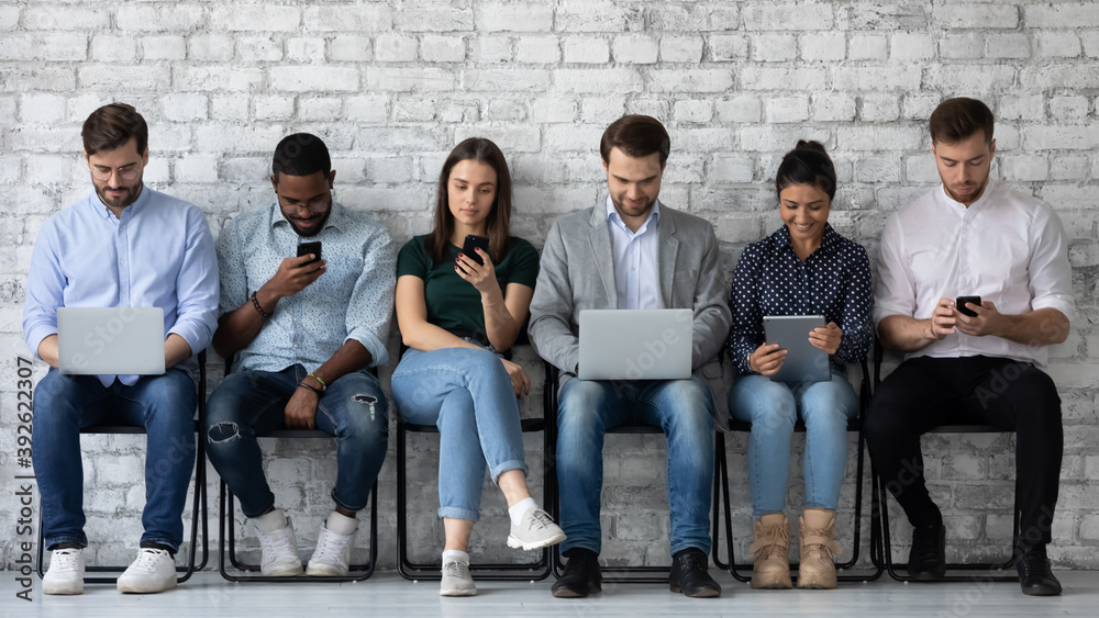 Modern way to communicate. Row of diverse young people sitting close to wall holding different gadgets cellphones laptops pads surfing internet chatting browsing social networks work or study online - obrazy, fototapety, plakaty 