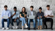 Modern way to communicate. Row of diverse young people sitting close to wall holding different gadgets cellphones laptops pads surfing internet chatting browsing social networks work or study online