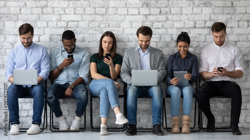 Obraz na płótnie Modern way to communicate. Row of diverse young people sitting close to wall holding different gadgets cellphones laptops pads surfing internet chatting browsing social networks work or study online