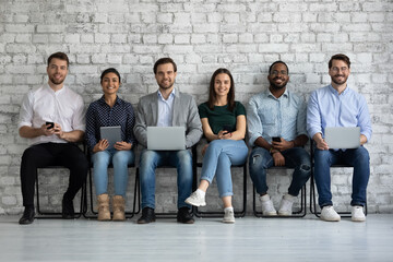 Portrait of young happy people of different gender and ethnicity job applicants business team group of clients posing on chairs in queue with diverse modern gadgets in hands smiling looking at camera