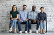 © fizkes - Patient waiting. Group portrait of four diverse people young men and women of different races sitting on chairs along the wall looking at camera expecting turn to visit doctor pass exam job interview