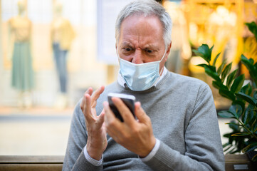 Senior man sitting on a bench and using a smartphone in a mall wearing mask and getting angry, coronavirus concept