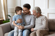 © fizkes - Happy three generations of men hugging, sitting on cozy couch at home, cute adorable little boy cuddling with young father and mature grandfather on sofa, family spending leisure time together