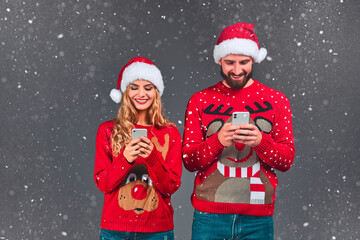 Married lovers are browsing, typing, reading at their phone, standing front the camera, wearing Christmas costumes.