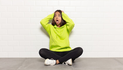 hispanic woman with open mouth, looking horrified and shocked because of a terrible mistake, raising hands to head