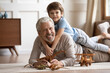 Portrait little grandchild lying on happy mature grandfather back, looking at camera, overjoyed smiling older man and adorable boy grandson hugging, having fun, lying on warm floor at home