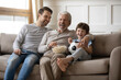 © fizkes - Laughing little boy with grandfather and father watching tv, eating popcorn snack, sitting on couch with soccer ball, happy family watching football game, spending leisure time at home together
