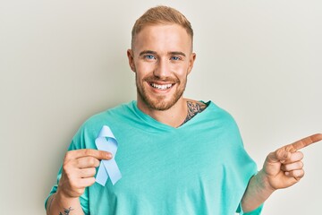 Young caucasian man holding blue ribbon smiling happy pointing with hand and finger to the side