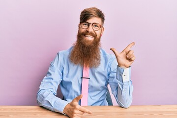 Wall Mural - Young irish redhead man wearing business shirt and tie sitting on the table cheerful with a smile on face pointing with hand and finger up to the side with happy and natural expression