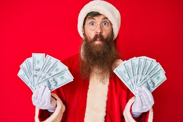 Canvas Print - Handsome young red head man with long beard wearing santa claus costume holding bunch of dollars making fish face with mouth and squinting eyes, crazy and comical.