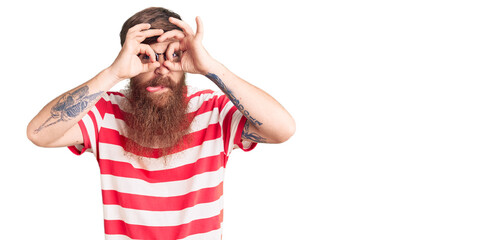 Wall Mural - Handsome young red head man with long beard wearing casual clothes doing ok gesture like binoculars sticking tongue out, eyes looking through fingers. crazy expression.