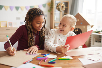 Portrait of two laughing teenage girls enjoying crafting and painting toether while siting at desk in decorated playroom