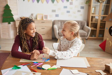 Portrait of two laughing teenage girls enjoying crafting and painting toether while siting at desk in decorated playroom, copy space