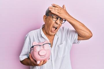 Canvas Print - Handsome mature senior man holding piggy bank with glasses stressed and frustrated with hand on head, surprised and angry face