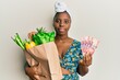 Young african woman holding groceries and 50 rand banknotes relaxed with serious expression on face. simple and natural looking at the camera.
