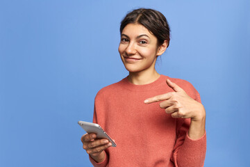 Pretty girl with nose piercing posing isolated holding smart phone, pointing fore finger at screen and smiling at camera, enjoying her new electronic gadget. Positive young woman messaging online