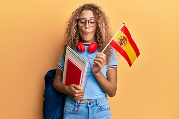 Sticker - Beautiful caucasian teenager girl exchange student holding spanish flag making fish face with mouth and squinting eyes, crazy and comical.