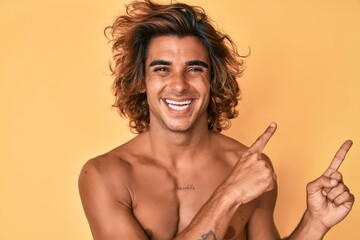 Young hispanic man standing shirtless smiling and looking at the camera pointing with two hands and fingers to the side.