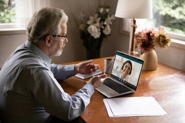 Side view focused elderly 60s businessman talking online with indian ethnicity female colleague, using computer video call application, consulting with helpline specialist, distant communication.