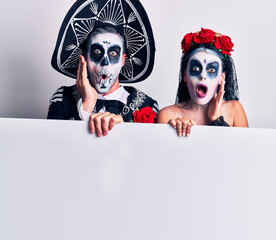 Wall Mural - Young couple wearing mexican day of the dead costume holding blank empty banner afraid and shocked, surprise and amazed expression with hands on face