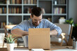 © fizkes - Smiling man sitting at workplace home office desk wearing glasses unpacking opening cardboard box long awaited parcel with goods bought on-line. Satisfied client of shipping delivery service concept