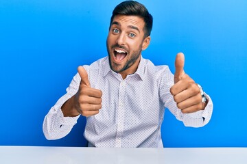 Handsome hispanic man wearing business clothes sitting on the table approving doing positive gesture with hand, thumbs up smiling and happy for success. winner gesture.