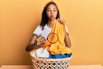 Wall Mural - Young african american girl holding magnifying glass looking for stain at clothes puffing cheeks with funny face. mouth inflated with air, catching air.