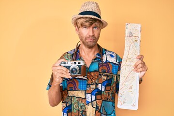 Poster - Young blond man holding vintage camera and city map relaxed with serious expression on face. simple and natural looking at the camera.