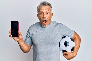 Sticker - Middle age grey-haired man holding football ball showing smartphone afraid and shocked with surprise and amazed expression, fear and excited face.