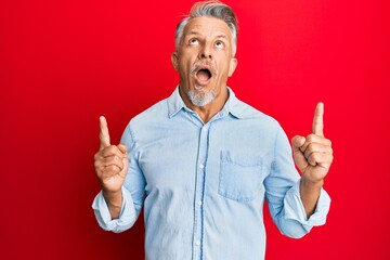 Middle age grey-haired man wearing casual clothes amazed and surprised looking up and pointing with fingers and raised arms.