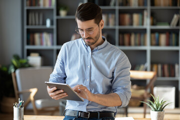 Focused serious businessman freelancer in glasses standing in modern office lean on table hold tablet device texting message to client, web surfing on-line, makes business offer to partner by e-mail