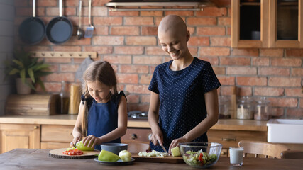 Our house speciality. Concentrated millennial mother recovering from cancer and little school age daughter enjoying cooking together at home kitchen, cutting ingredients for vegetable dish on lunch