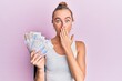 Beautiful blonde sport woman holding 20 swedish krona banknotes covering mouth with hand, shocked and afraid for mistake. surprised expression