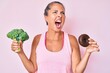 © Krakenimages.com - Middle age hispanic woman holding broccoli and chocolate donut angry and mad screaming frustrated and furious, shouting with anger. rage and aggressive concept.