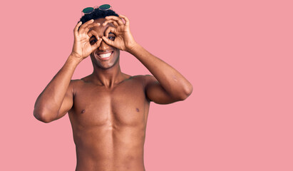 Wall Mural - African handsome man wearing swimsuit and sunglasses doing ok gesture like binoculars sticking tongue out, eyes looking through fingers. crazy expression.