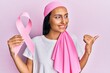 © Krakenimages.com - Young brunette woman wearing head scarf and holding pink cancer ribbon pointing thumb up to the side smiling happy with open mouth