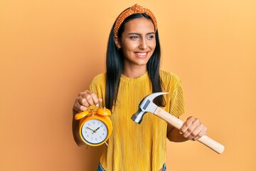 Canvas Print - Young brunette woman holding alarm clock and hammer smiling looking to the side and staring away thinking.