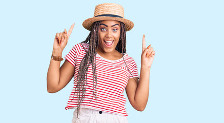 Canvas Print - Young african american woman with braids wearing summer hat smiling amazed and surprised and pointing up with fingers and raised arms.