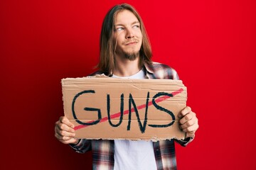 Handsome caucasian man with long hair holding no guns warning banner smiling looking to the side and staring away thinking.